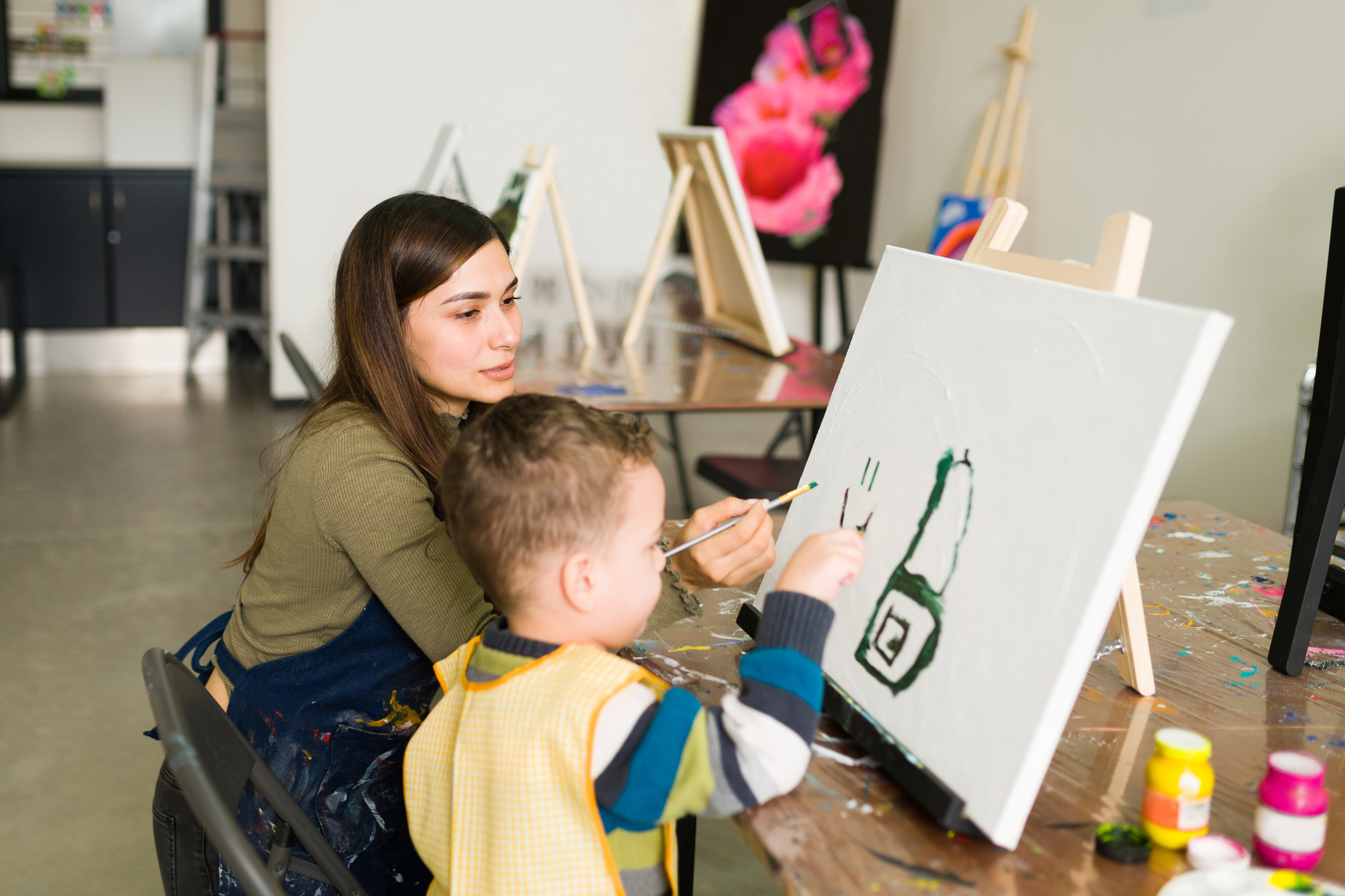 Young teacher and a kid student with aprons are painting on a canvas